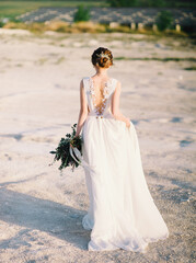 Fototapeta na wymiar Desert wedding, sunset, golden hour time. Yong bride in white lace dress goes in the desert and keeping wedding bouquet with tender flowers.
