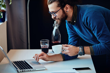 Young attractive caucasian hipster standing in his office, holding cup of coffee and checking on e-mail from his boss.