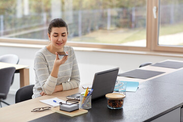 remote job, business and people concept - smiling young woman with smartphone and laptop computer using voice command recorder on smartphone at home office