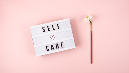 Self-care word on lightbox and flower narcissus on pink background flat lay. Take care of yourself