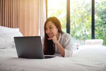 A beautiful asian woman using and working on laptop computer while lying down on a white cozy bed at home