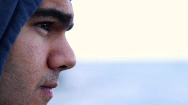Caucasian man wearing hoodie and coat gazes out on sea water, close up profile