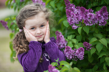 Cute toddler girl with loose curly hair among the bushes of lilac with bunches of flowers
