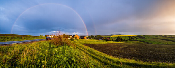 Spring rapeseed and small farmlands fields after rain evening view, cloudy pre sunset sky with colorful rainbow and rural hills. Natural seasonal, climate, farming, countryside beauty concept scene.