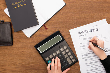 Fototapeta na wymiar Partial view of woman filling in bankruptcy form and using calculator on wooden background