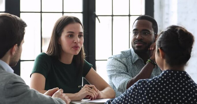 Young female team leader sitting at table with mixed race colleagues, discussing working issues at negotiations meeting. Skilled millennial diverse employees developing startup marketing strategy.