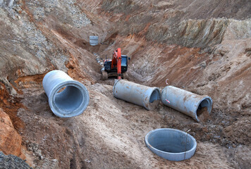 Excavator working at construction site on earthworks. Backhoe digs ground in pit for laying concrete sewer wells and sewage pipes. Installing underground storm systems of water main and sanitary sewer