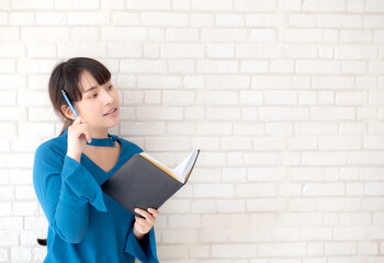 Beautiful asian woman smiling standing thinking and writing notebook on concrete cement white background at home, girl homework on book, education and lifestyle concept.