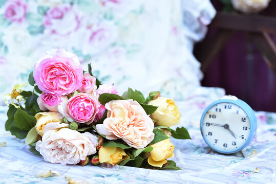 Cute bouquet with colorful roses on the bed and an alarm clock. Photo in vintage style, provence. Cozy rest in the garden.