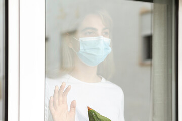 Day of quarantine for a young woman with a face blue mask looking out a window resting his hand on the glass melancholic