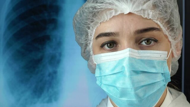 Portrait of medic woman on background of X-ray of lungs with pneumonia. Doctor looks at camera and corrects medical mask hands in medical gloves. Close-up. Coronavirus. COVID-19.