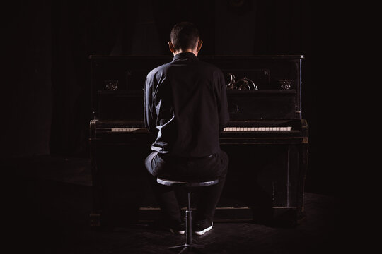 Man in black suit sit in front of piano in black interior, rear view.