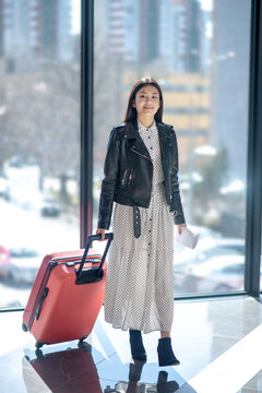 Brunette female in leather jacker walking with red suitcase, smiling