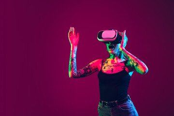 Playing with VR-headset. Young caucasian woman on purple studio background in neon light. Beautiful female model with tattoos. Human emotions, facial expression, sales, ad concept. Freak's culture.