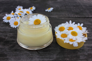 Natural chamomile face cream and fresh daisy flowers on dark rustic wooden background. Homemade beauty product. Organic cosmetic and self-care concept. Top view, copy space.
