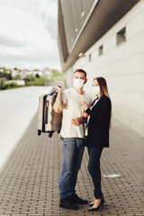 Young man and woman in protective masks standing near the airport. Man holding a suitcase. Safe travel after a coronavirus pandemic. Happy young couple flying on a journey after quarantine