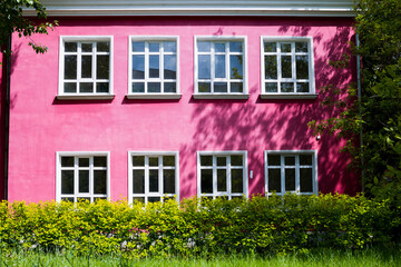 Fototapeta na wymiar A bright pink building with large Windows surrounded by bright green trees and shrubs