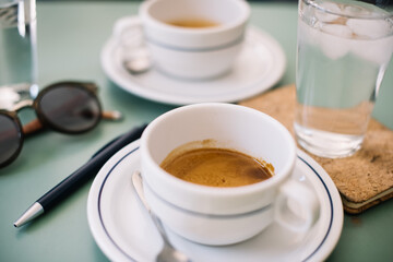 Two cups of delicious hot espresso coffee  on the green table background, standing along with glass of ice cold water, pen, paper notebook and sunglasses