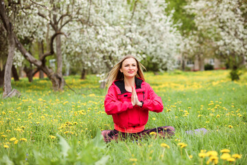 Naklejka na ściany i meble Beautiful woman is practicing yoga outdoors in the blooming park, holding hands in namaste. Woman is dressed in pink bomber jacket. 