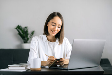 Woman using laptop at workplace at home office and writing notes in notepad. Frelance work from home in quarantine concept.