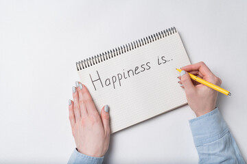 Copybook with HAPPINESS IS text. Top view of female hands writing in Notebook