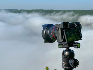 A camera with a tripod at sunrise looks at a foggy canyon.