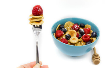 A metal fork with Stack of mini tiny pancakes and berry near blue bowl with heap of cereal mini pancakes with honey spoon. Trend 2020. Home made long breakfast during quarantine. White background. 