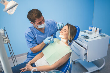 Obraz premium Dentist examining a patient's teeth using dental equipment in dentistry office. Stomatology and health care concept. Young handsome male doctor in disposable medical facial mask, smiling happy woman.