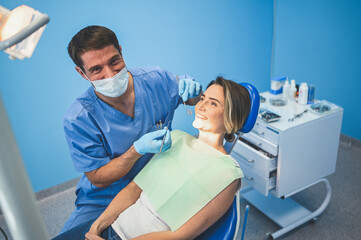 Fototapeta na wymiar Dentist examining a patient's teeth using dental equipment in dentistry office. Stomatology and health care concept. Young handsome male doctor in disposable medical facial mask, smiling happy woman.