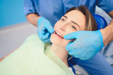 Dentist examining a patient's teeth using dental equipment in dentistry office. Stomatology and health care concept. Young handsome male doctor in disposable medical facial mask, smiling happy woman.