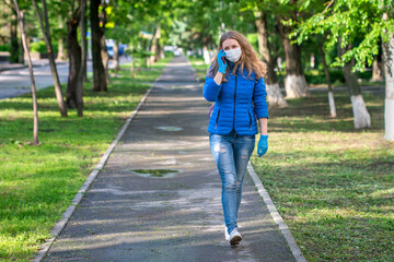 Fototapeta premium A woman in protective mask and gloves is walking along empty street and talking on phone. Lifestyle and safe behavior during a coronavirus pandemic. The new normal and modern reality. Copy space.