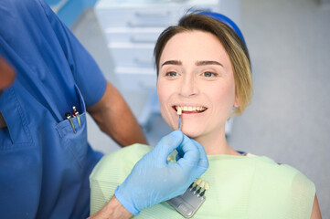Dentist checks the level of patient's teeth whitening with a dentist's color. Dental equipment in dentistry office. Stomatology concept. Doctor's hands in medical gloves and smiling happy woman.