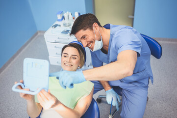 Fototapeta premium Dentist shows the patient the results of treatment with a mirror, examinating teeth with dental equipment in dental office. Young handsome male doctor in medical facial mask and smiling happy woman.