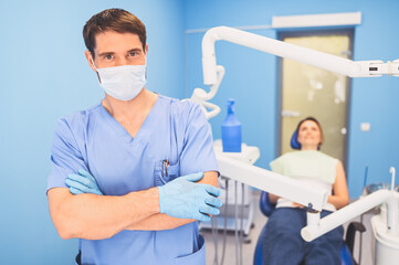 Young smiling handsome male doctor in blue medical uniform, disposable medical facial mask with equipment in dentistry office and happy patient in the dental chair. Stomatology concept. Copy space