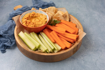 Bowl of butternut squash hummus on a wooden tray with carrots, celery, and pita bread against a blue textured background