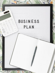 Business Plan text on letter board and open notebook with pen and calculator on money hundred dollar bills background. Business plan poster or web banner. Small business how to start poster