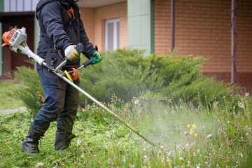 A man with a lawn mower on a grassy field near the house. A man in protective clothing, gloves and rubber boots mows the grass with a trimmer.