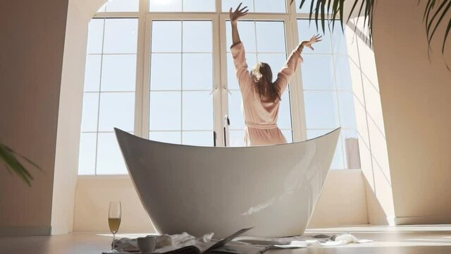 A young woman with her arms outstretched is sitting on the bathtub after a night's sleep on a Sunny morning