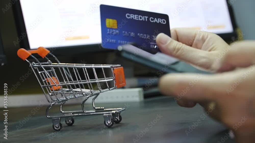 Poster close up Shallow focus hand hold Credit Card and touch mobile phone screen with small shopping cart and E-commerce website on monitor background. Background for E-commerce and online shopping concept.