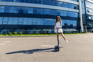 A girl in a dress rides an electric scooter near modern buildings