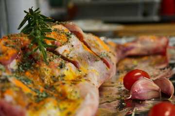 Raw leg of lamb on a tray with aluminum foil. Rosemary, garlic, chery tomato, salt, cumin, oregano,...