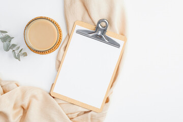 Modern home office desk with clipboard mockup, cup of coffee, beige blanket and eucalyptus flower on white background. Freelancer workspace concept. Flat lay, top view.