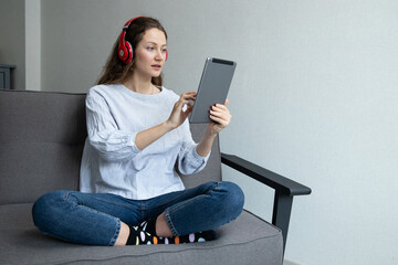 A girl in red headphones is typing on a tablet sitting at home on the couch