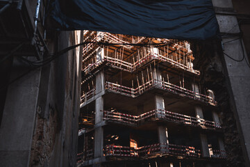 A contrast view through a concrete hole of a construction site: an unfinished high-rise building with bare walls and plenty of temporary wooden fences with warning tape on them on each storey