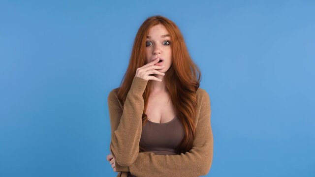 Red-haired lady in brown t-shirt and cardigan. She is smiling and looking surprised, saying wow while posing against blue studio background. Close up