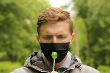 Masked man in the park. Quarantine problem. A man cannot eat a lollipop. Discomfort due to facial mask. Black mask guy
