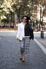 Naklejka premium Autumn fashion outdoor. The girl in fashionable stylish white shirt , pants, jacket and sunglasses, autumnal lifestyle on the background of blurry yellow-green trees in the park. Vertical