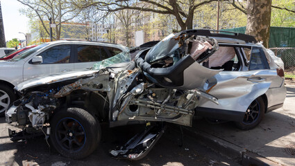 a totaled, abandoned car half on the sidewalk, half on the street. Silver color body with the exploded airbag visible through the smashed window