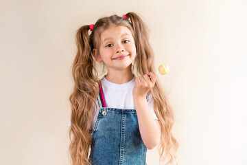A little girl with long hair stands in a denim jumpsuit and holds a round Lollipop in her hand.