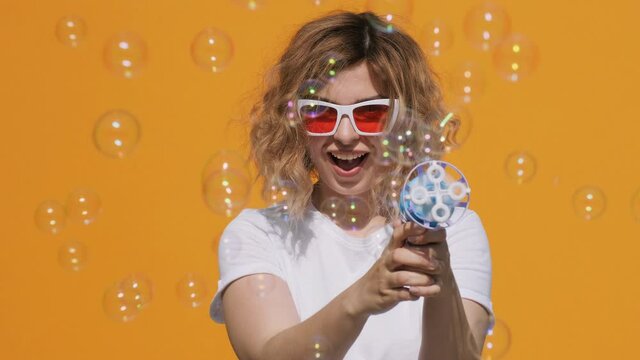 Portrait of happy woman in stylish sunglasses with soap bubbles smiling releases toys from gun with pleasure on yellow background in summer slow motion. Emotions, joy. Positive flirty girl. Childhood